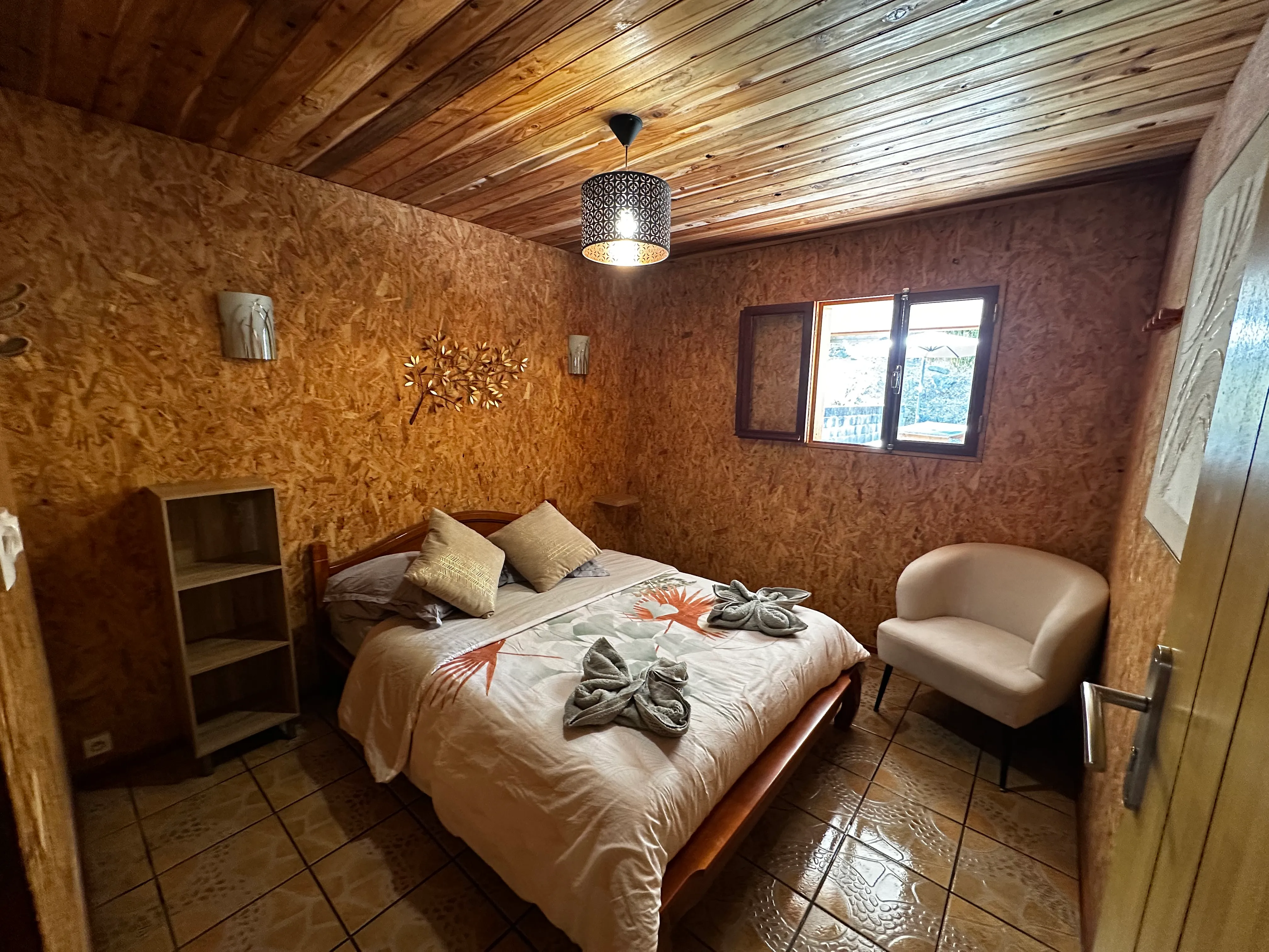 Chambre double n°1 du gîte : lit double avec coussins, murs et plafond en bois, fauteuil clair